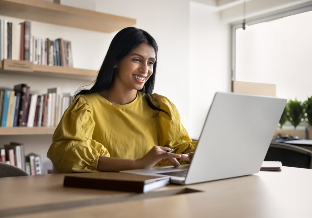 Cheerful successful young Indian project manager woman enjoying online communication Cheerful successful young Indian project manager woman enjoying online communication at computer, typing on laptop in co-working space, smiling, working, using job application, browsing Internet